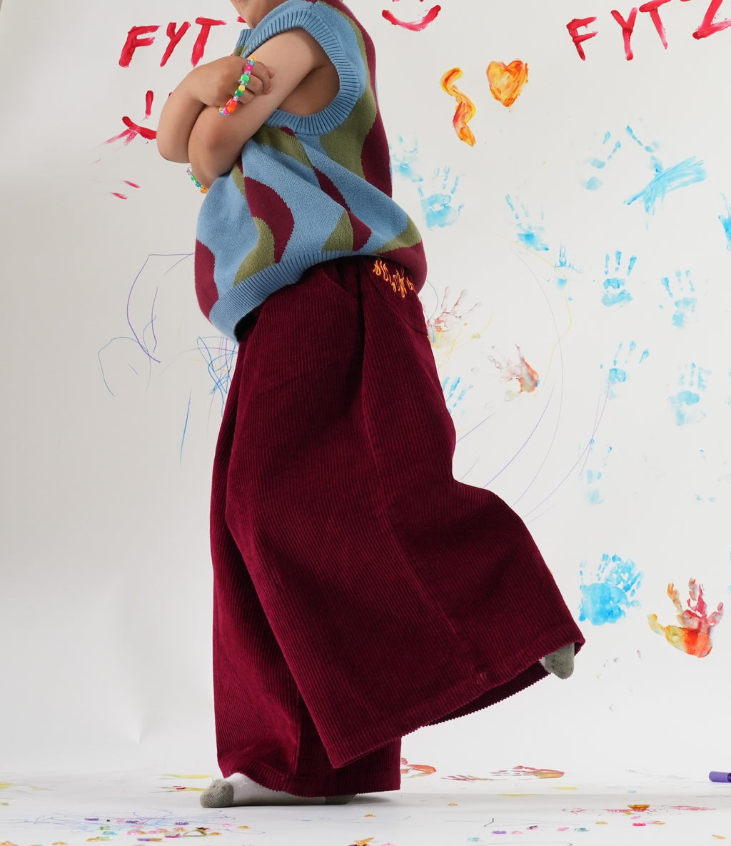 Child wearing a colorful Johnny vest in cherry ice swirl and red wide-leg Blaze cord pants in front of a wall with child's drawings.