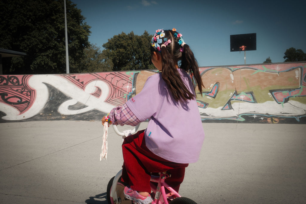 Child on a bicycle in a skate park with graffiti on the wall wearing Ollie layered double-sleeve shirt in fig purple tone — From Y To Z