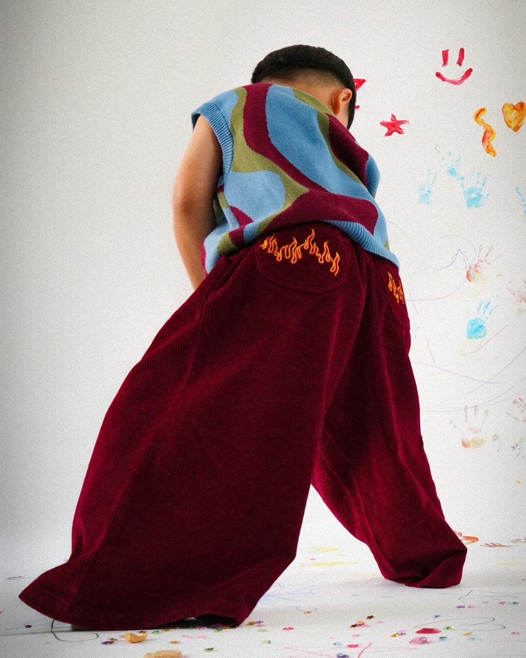 Person wearing a colorful top and maroon corduroy flame blaze pants on a white background with colorful footprints.