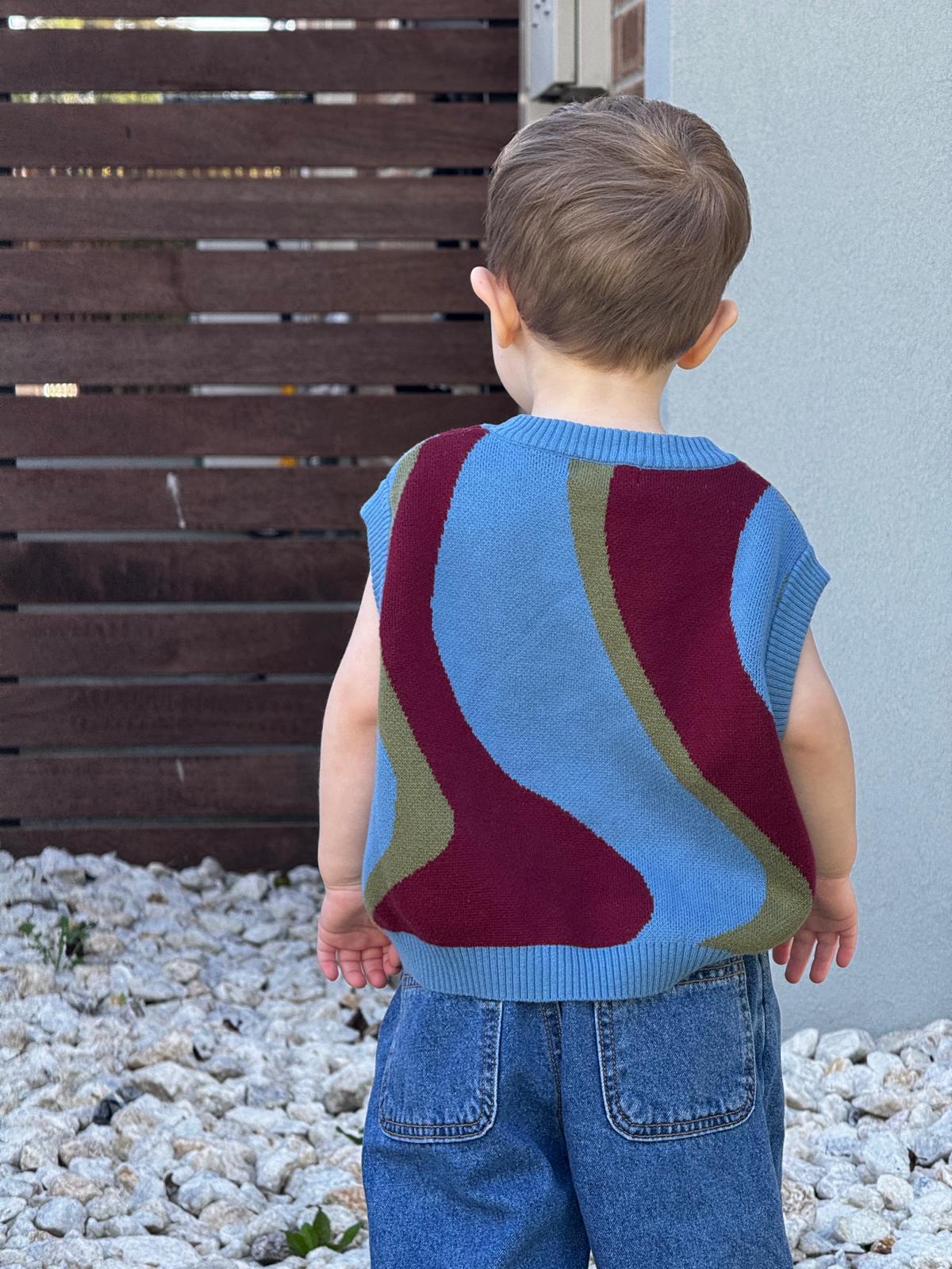 Child wearing a colorful sleeveless johnny sweater vest standing against a wooden fence and gray wall.
