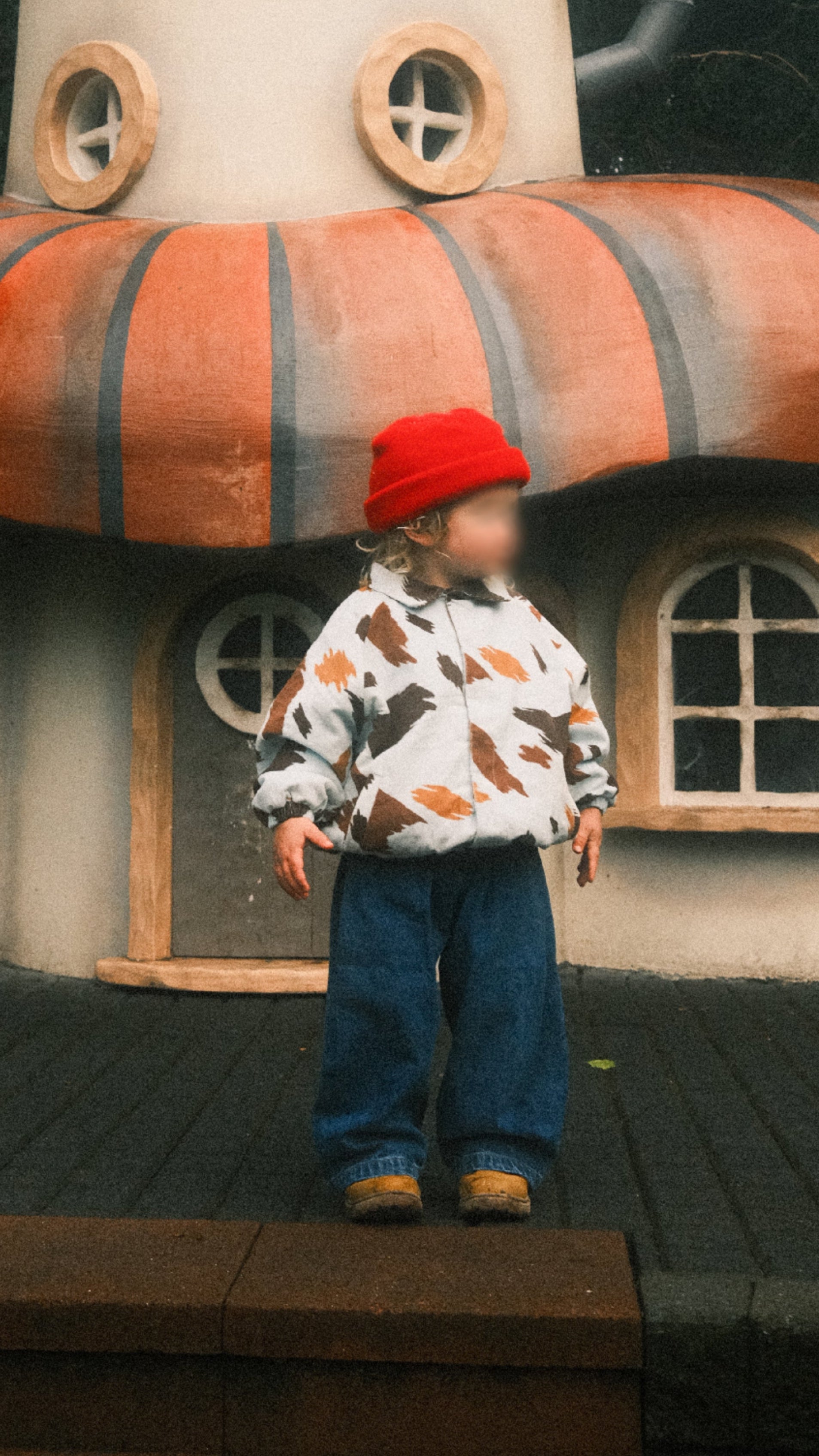 A child wearing a red beanie, seffi jacket jacket, and ruhi blue jeans stands in front of a whimsical mushroom-shaped house with orange and dark stripes.