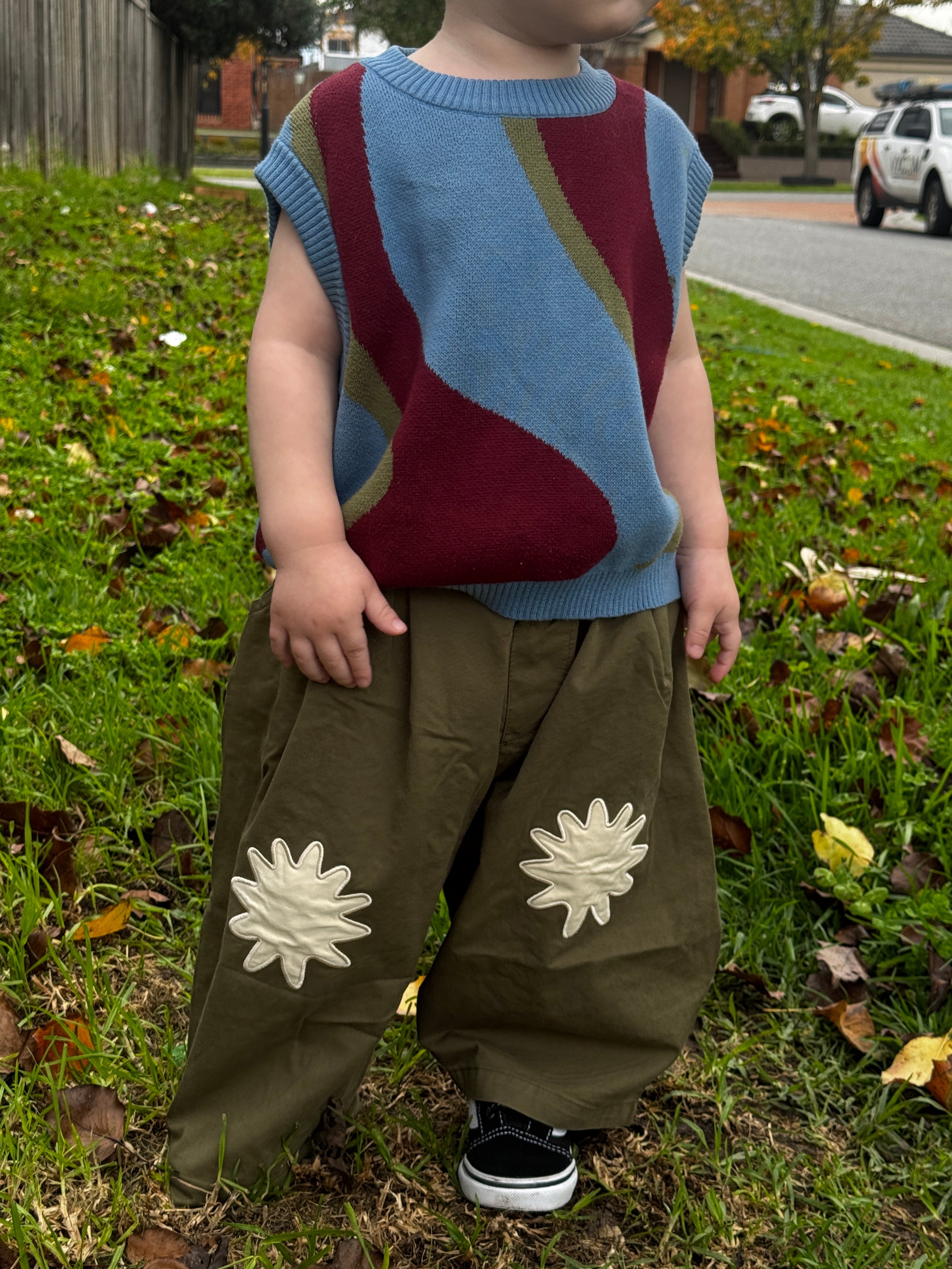 Child wearing a colorful johhny vest and green pants with white flower patterns on grass.
