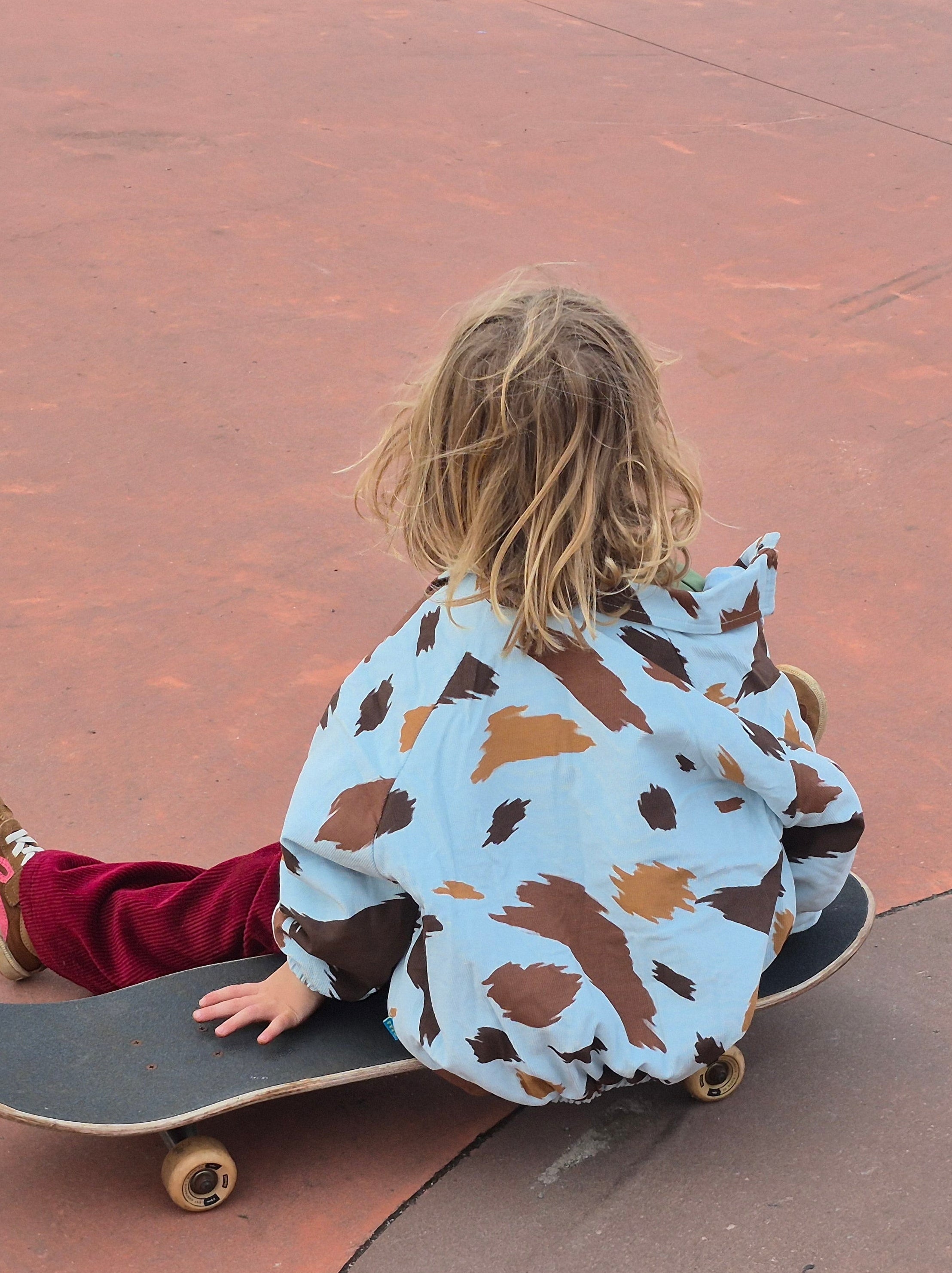 Child sitting on a skateboard wearing a seffi cow print patterned jacket in an outdoor setting