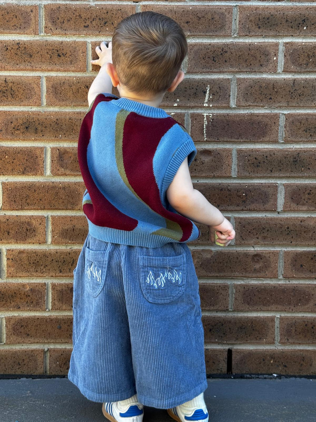 Child wearing a colorful johnny sweater vest and blue pants against a brick wall.