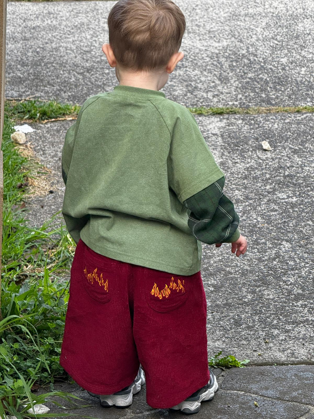 Child wearing a green plaid long sleeve and red corduroy pants with flame embroidery, standing on a sidewalk.